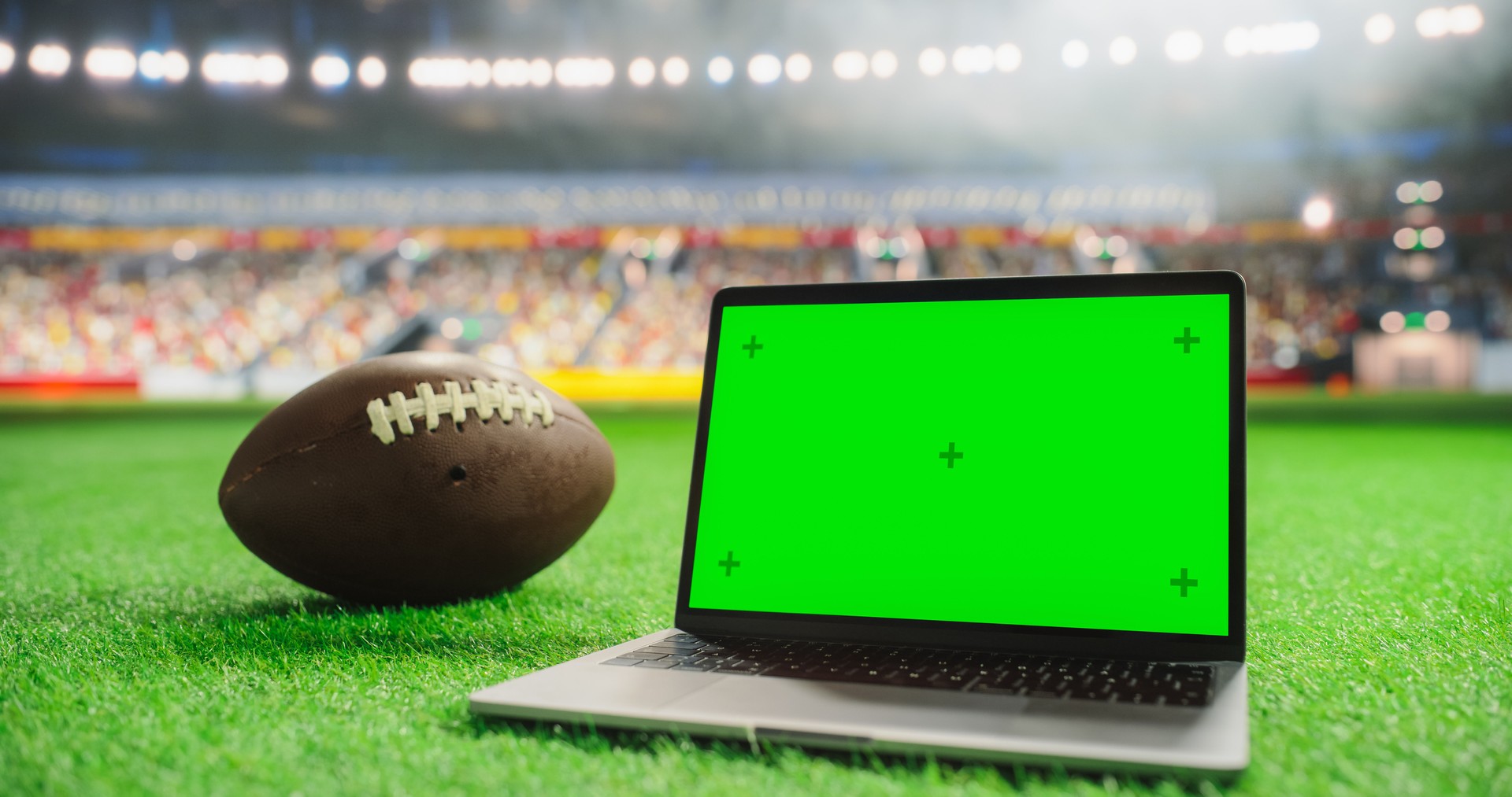 Laptop Computer with a Green Screen Mock-Up Display Positioned Next to American Football on a Stadium Pitch at Night. Template for Software, Streaming, Sports Betting, Multimedia Content